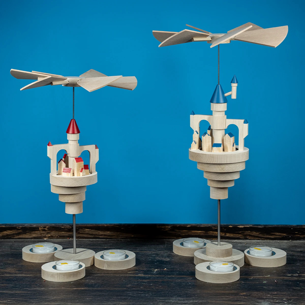 Zwei hölzerne Pyramiden mit Flügeln stehen auf einer Holzfläche Die eine hat rote und die andere blaue Türme mit kleinen Häusern darunter Beide haben Teelichthalter Im Hintergrund blauer Untergrund