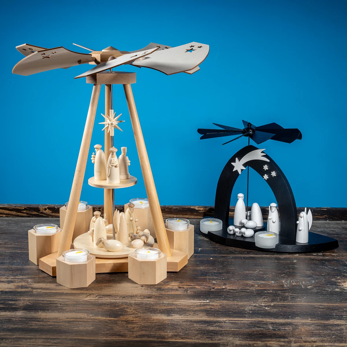 Eine hölzerne Pyramide mit Flügeln und Figuren sowie Kerzenhaltern steht auf einer Holzfläche Daneben eine schwarze Pyramide mit weißen Figuren und Sternen Im Hintergrund blauer Untergrund